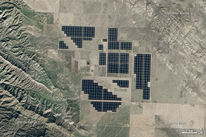 Satellite view of a large photovoltaic array arranged in geometric blocks across dry grassland.